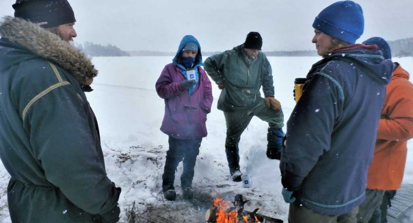 A group of people stand around a campfire in a snowy landscape. Some are holding mugs. 